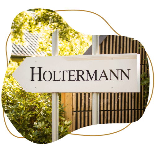 Brauner Klecks mit klecksförmigen Bild davor. Im Klecks ist ein pfeilförmiges Schild mit der Aufschrift 'Holtermann' zu lesen