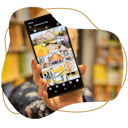 Brauner Klecks mit klecksförmigen Bild davor. Im Klecks ist eine Hand zu sehen, die ein Handy mit der Übersicht der Holtermann-Seite auf Instagram zum Betrachter streckt